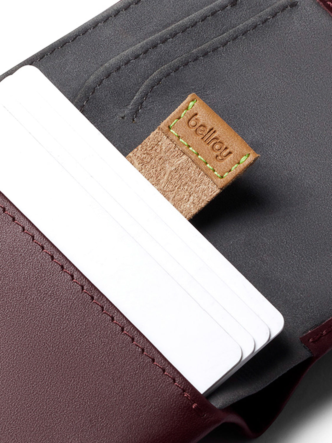 A close-up of the Bellroy Note Sleeve Wallet in maroon and gray leather, holding three white cards, features a brown tab with "Bellroy" and green stitching.