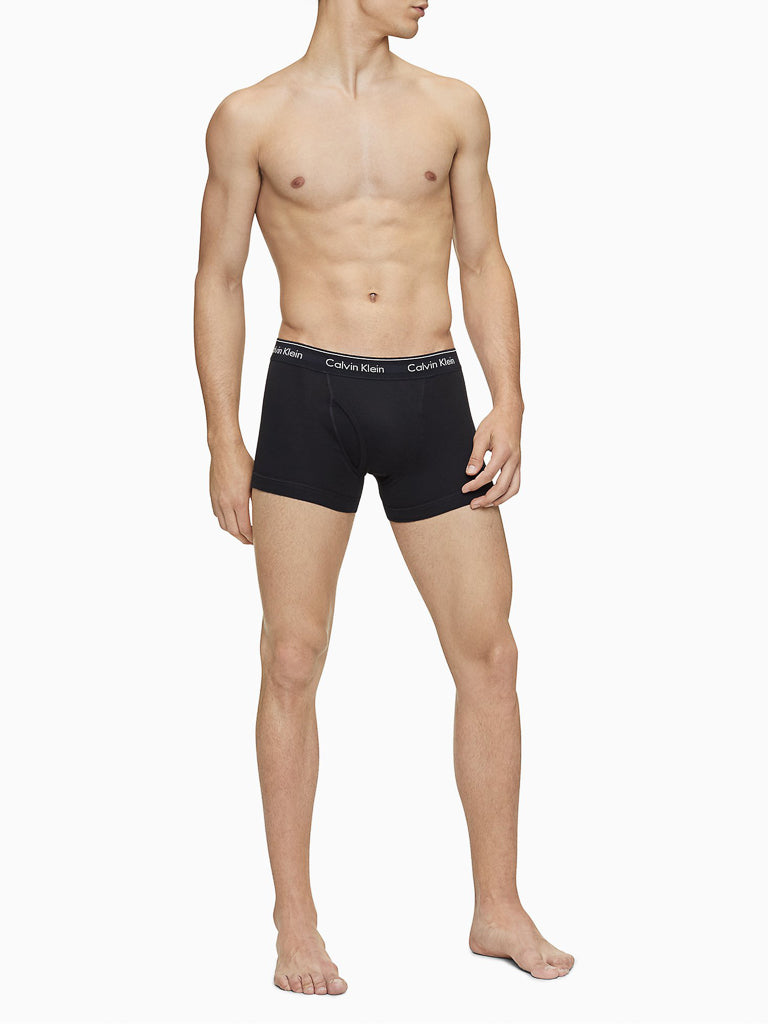A shirtless man models Calvin Klein NB4002 3-Pack Cotton Classic Fit Trunk underwear in black, standing barefoot against a plain white background and looking slightly to the side.