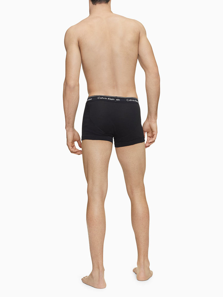 A shirtless man stands barefoot, facing away from the camera, wearing Calvin Klein NB4002 3-Pack Cotton Classic Fit Trunk men's underwear.