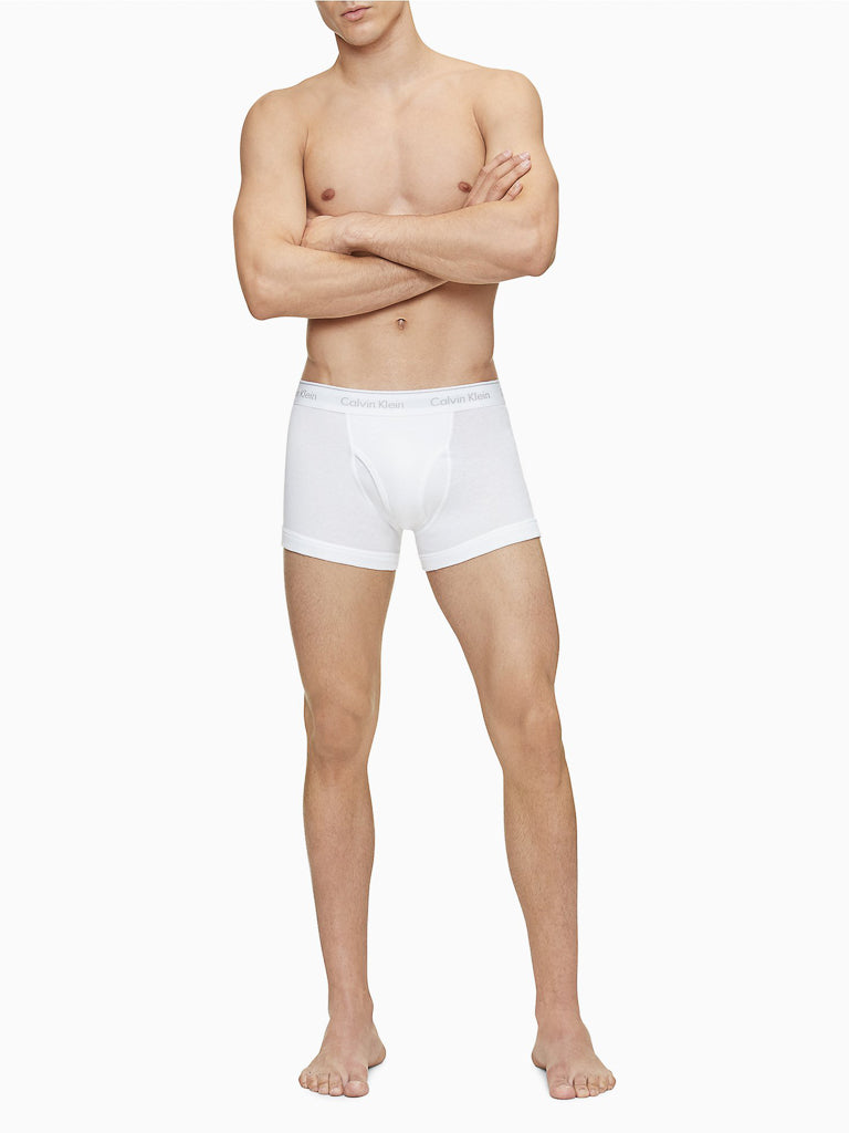 A man stands barefoot with arms crossed, shirtless, facing forward against a plain white background, wearing Calvin Klein NB4002 3-Pack Cotton Classic Fit Trunk in white.
