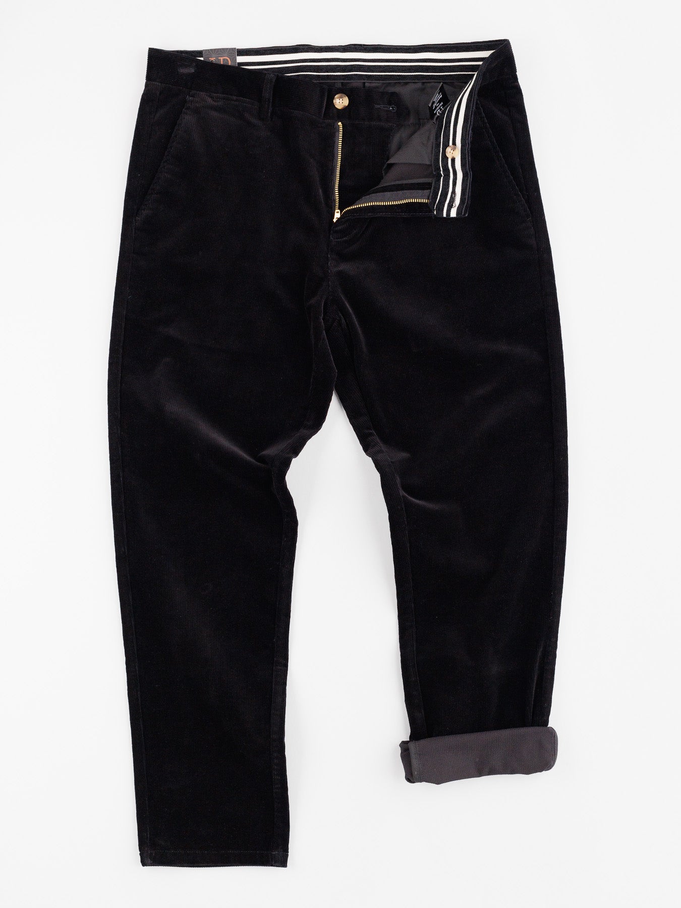 The Skillman Corduroy Pant by ID Menswear is displayed on a white background. It has a partially open zip fly, button closure, side pockets, one roll-up cuff on the right leg, a 32 inseam, and an interior waistband with a striped pattern.