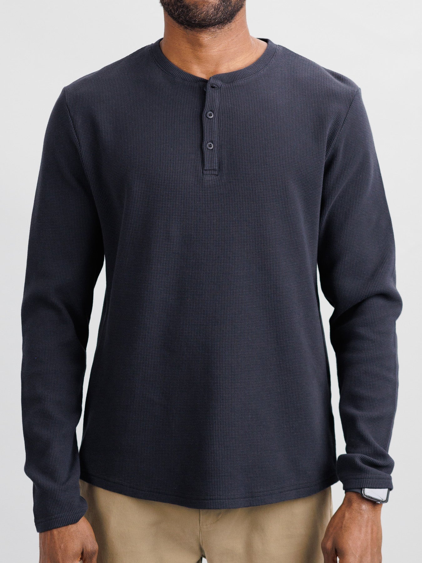 A person in ID Menswears Freestyle Honeycomb Knitted Mesh Henley in dark blue, featuring a textured pattern and buttons at the neckline, pairs it with khaki pants. Made from soft organic cotton, they have a short beard and are set against a plain background.