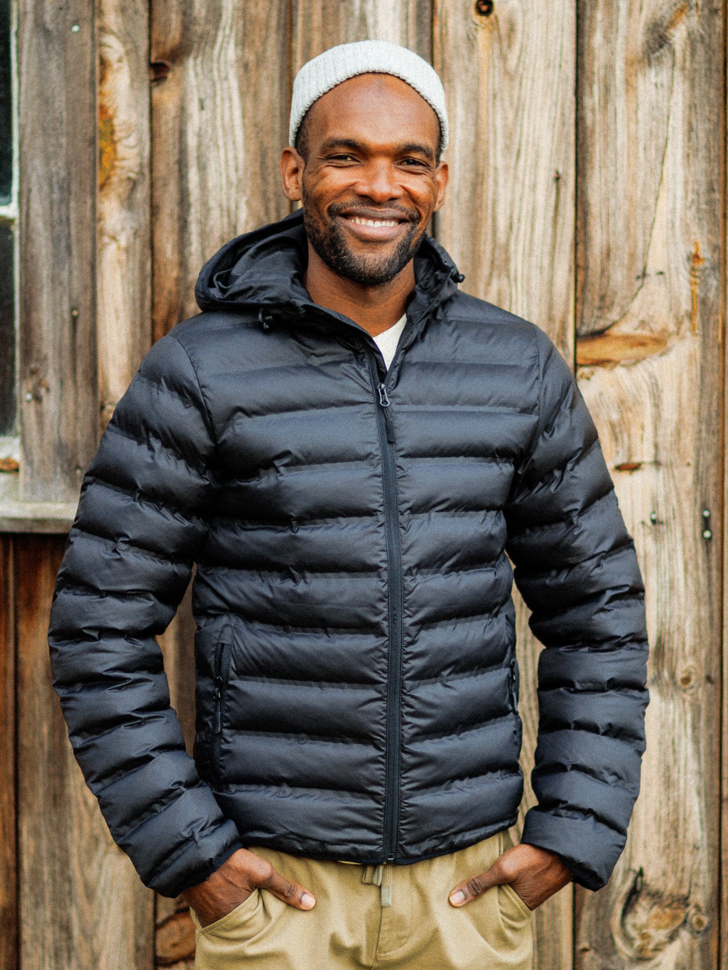 A smiling man in a gray beanie, beige pants, and the ID Menswear Yukon Hooded Puffer Jacket stands before a rustic wooden wall.
