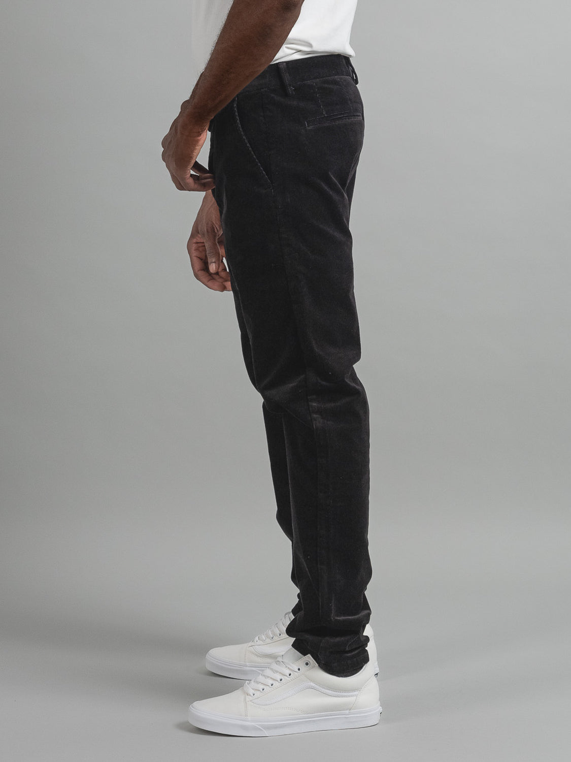 A person is seen in a side profile view wearing ID Menswears Skillman Corduroy Pants in black and white sneakers. The setting features a plain light gray background, with the persons upper body partly visible in a crisp white shirt.