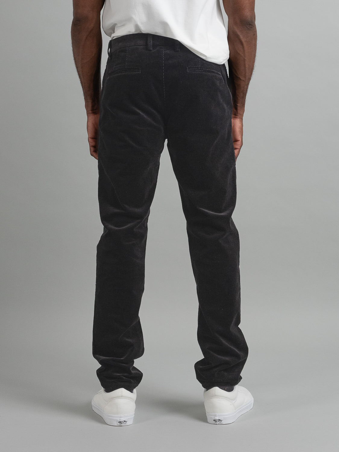 Facing away from the camera, a person effortlessly blends smart-casual style in ID Menswears Skillman Corduroy Pant with a 32 inseam and white sneakers, accented by a crisp white shirt against a plain light gray backdrop.