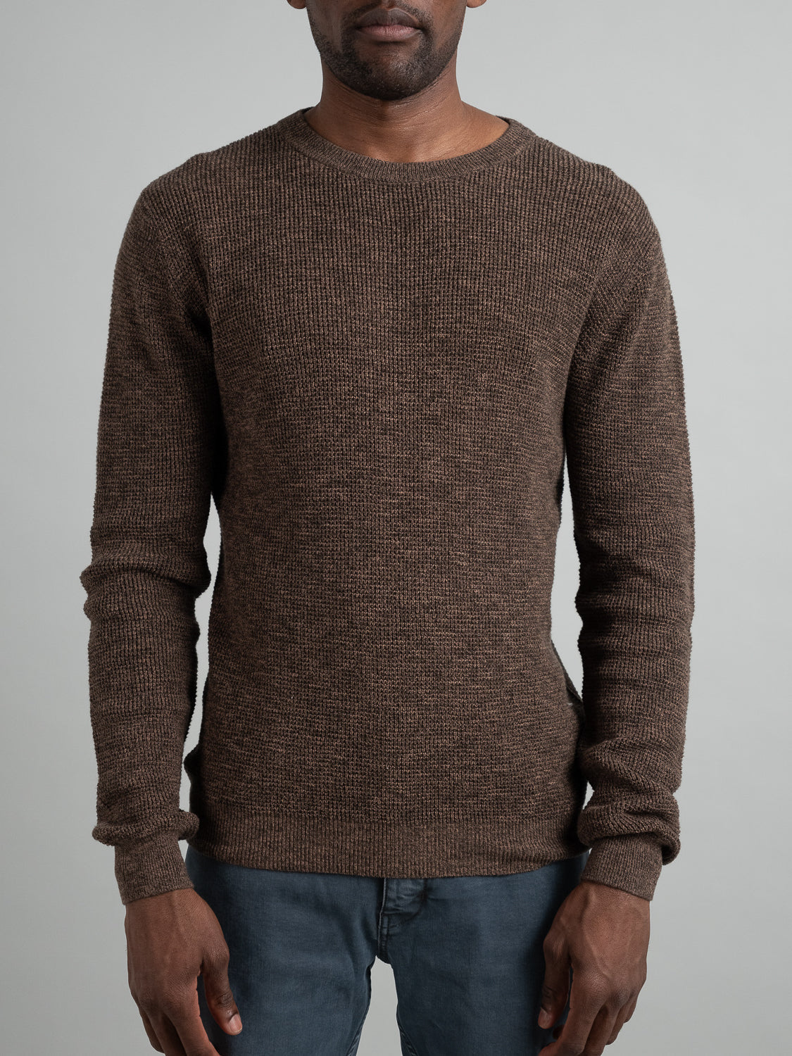 A person in an Oslo Waffle Knit Sweater by ID Menswear and dark jeans stands against a neutral background. The brown sweaters ribbed texture adds depth, with the individuals hands resting casually by their sides.