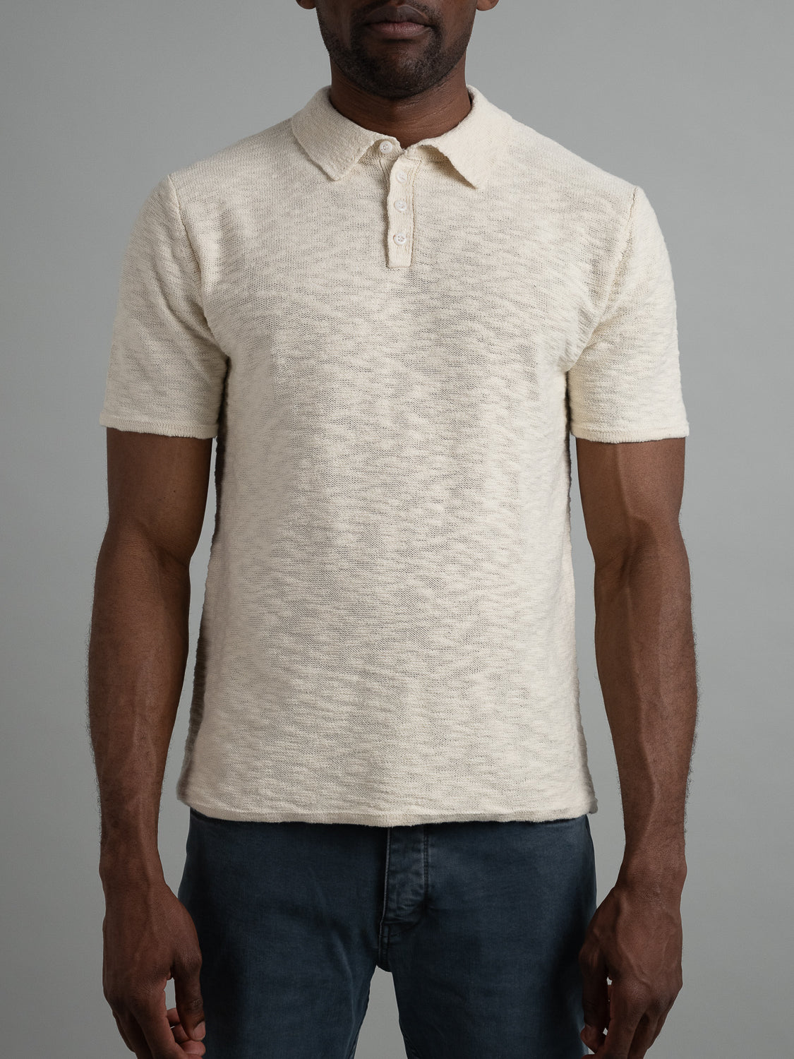 A person in a light beige Miami Reverse Knit Cotton Polo by ID Menswear and dark fitted jeans stands against a plain gray background. The breathable polo, featuring a collar and three buttons with a subtle pattern, pairs perfectly with the casual style of the jeans.
