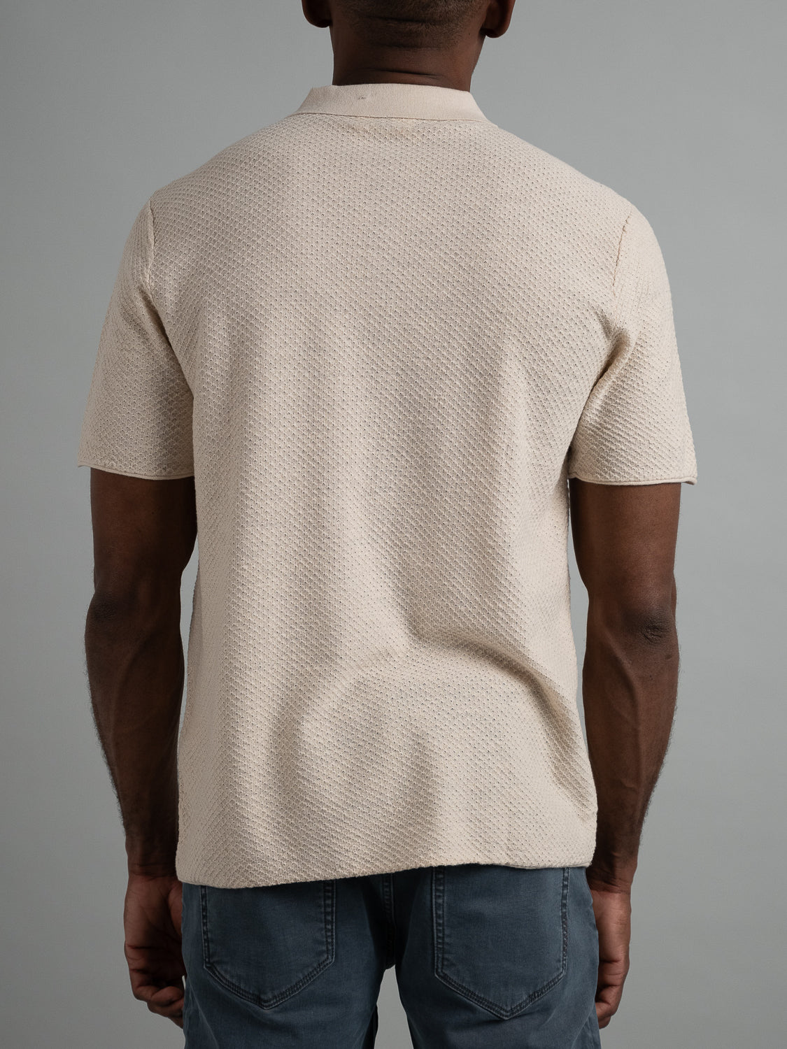 A person in the Phoenix Super Fine Cotton Knit Polo by ID Menswear and dark jeans stands facing away, showcasing a versatile look against a plain gray background.