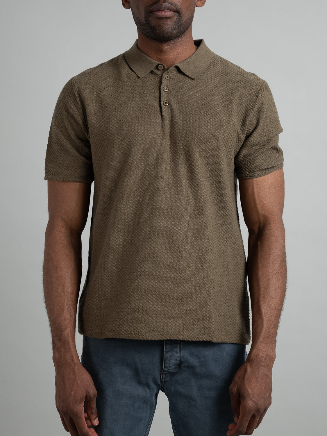 Against a gray backdrop, a person models the Phoenix Super Fine Cotton Knit Polo by ID Menswear in olive green. The textured shirt features a pointed collar and three buttons, paired with blue jeans, offering a versatile and stylish look as they stand facing forward with arms at their sides.