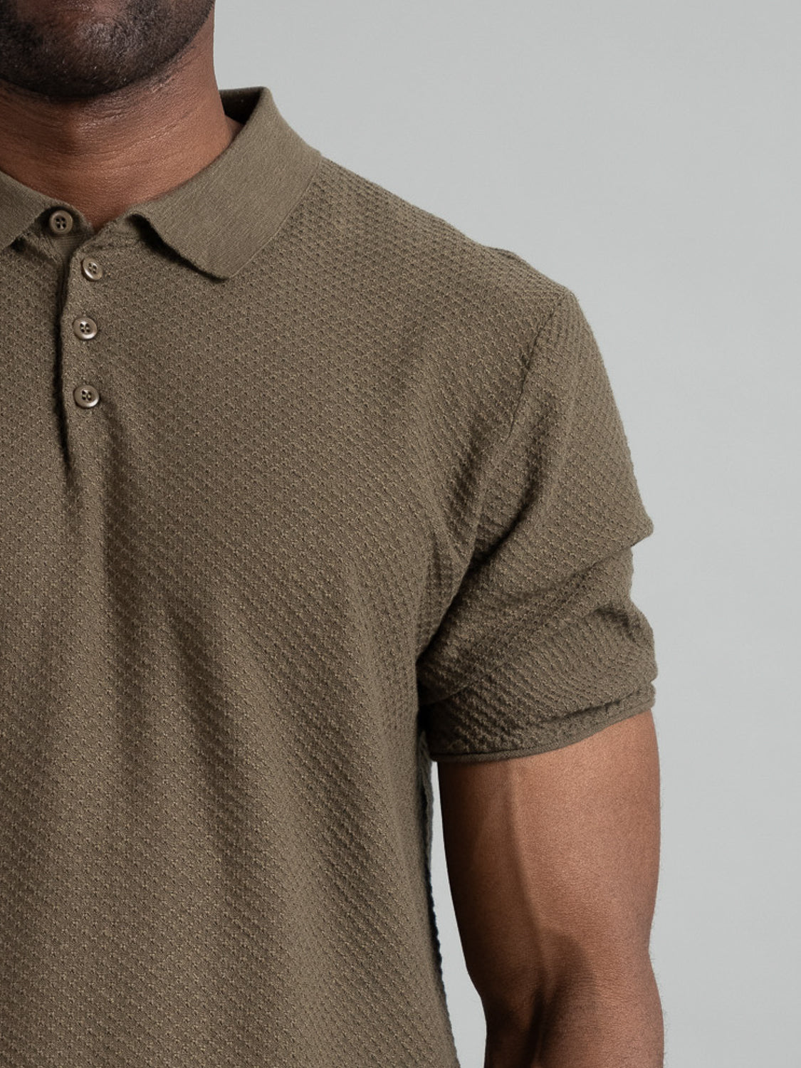 Close-up of a person in an olive green Phoenix Super Fine Cotton Knit Polo by ID Menswear, showcasing versatile style. The textured cotton-knit design features a buttoned collar, with the upper torso and part of the right arm against a neutral light gray backdrop.