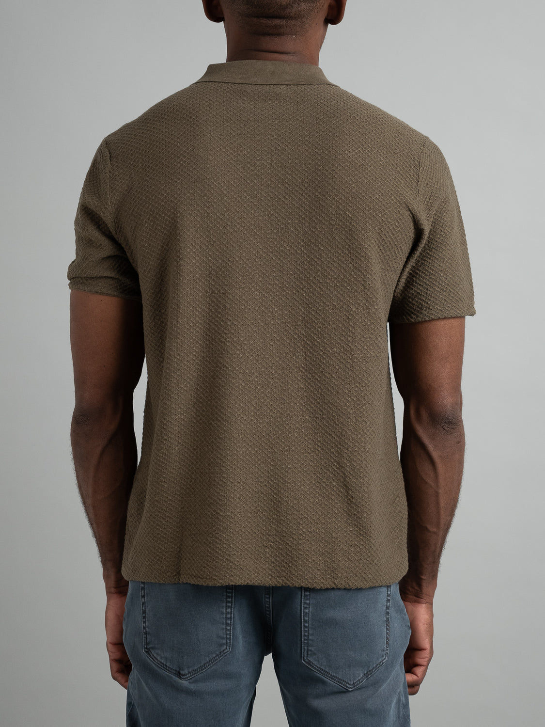 A person with short hair stands with their back to the camera, embodying a versatile look in an olive green Phoenix Super Fine Cotton Knit Polo by ID Menswear and blue jeans, ideal for casual wear.