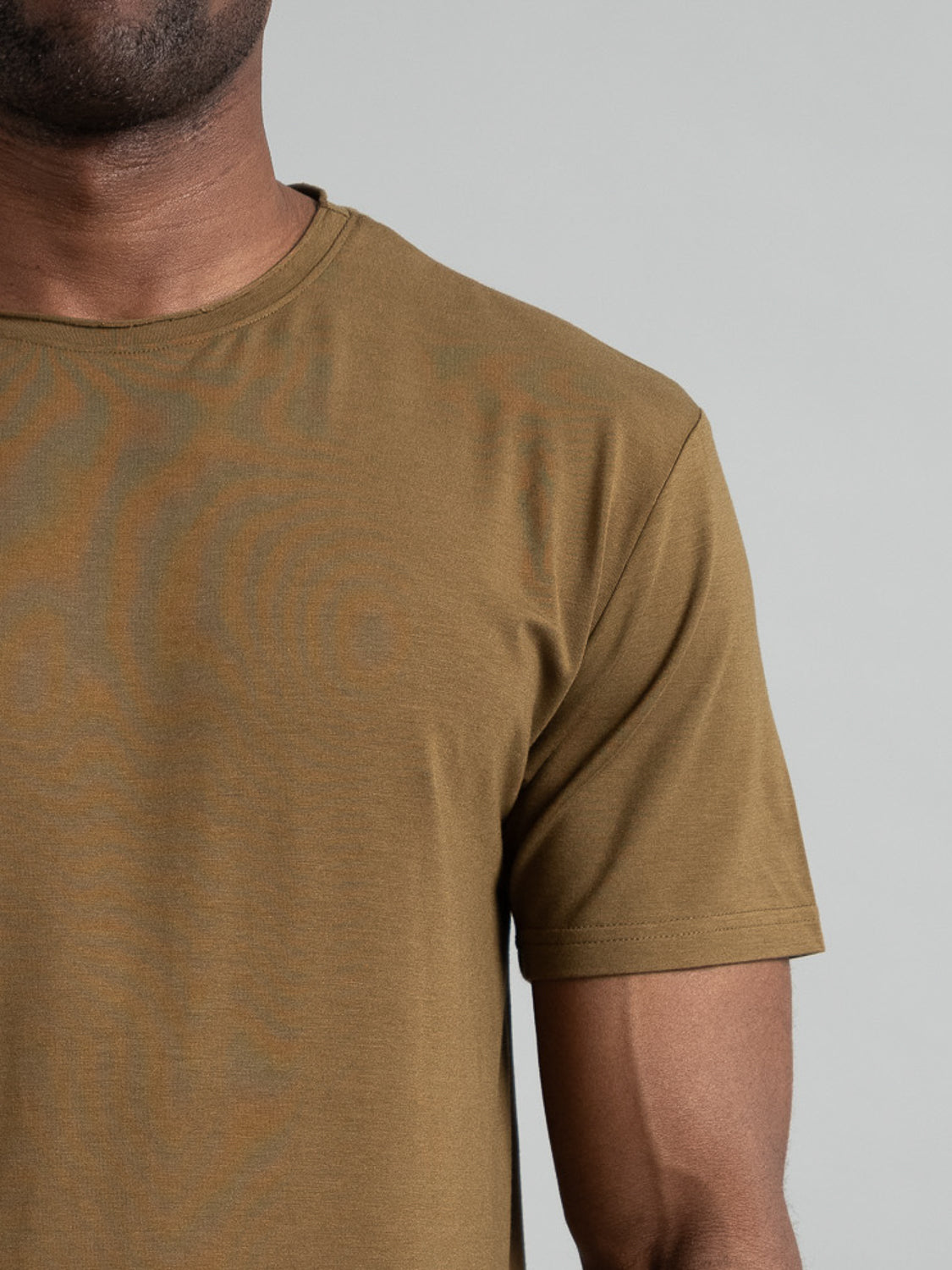 An individual sports the ID Menswear Bello Bamboo Blend Rolled Neck Tee in plain olive green, with a cropped view from neck to mid-arm, set against a muted light gray background highlighting the sustainable fashion choice.