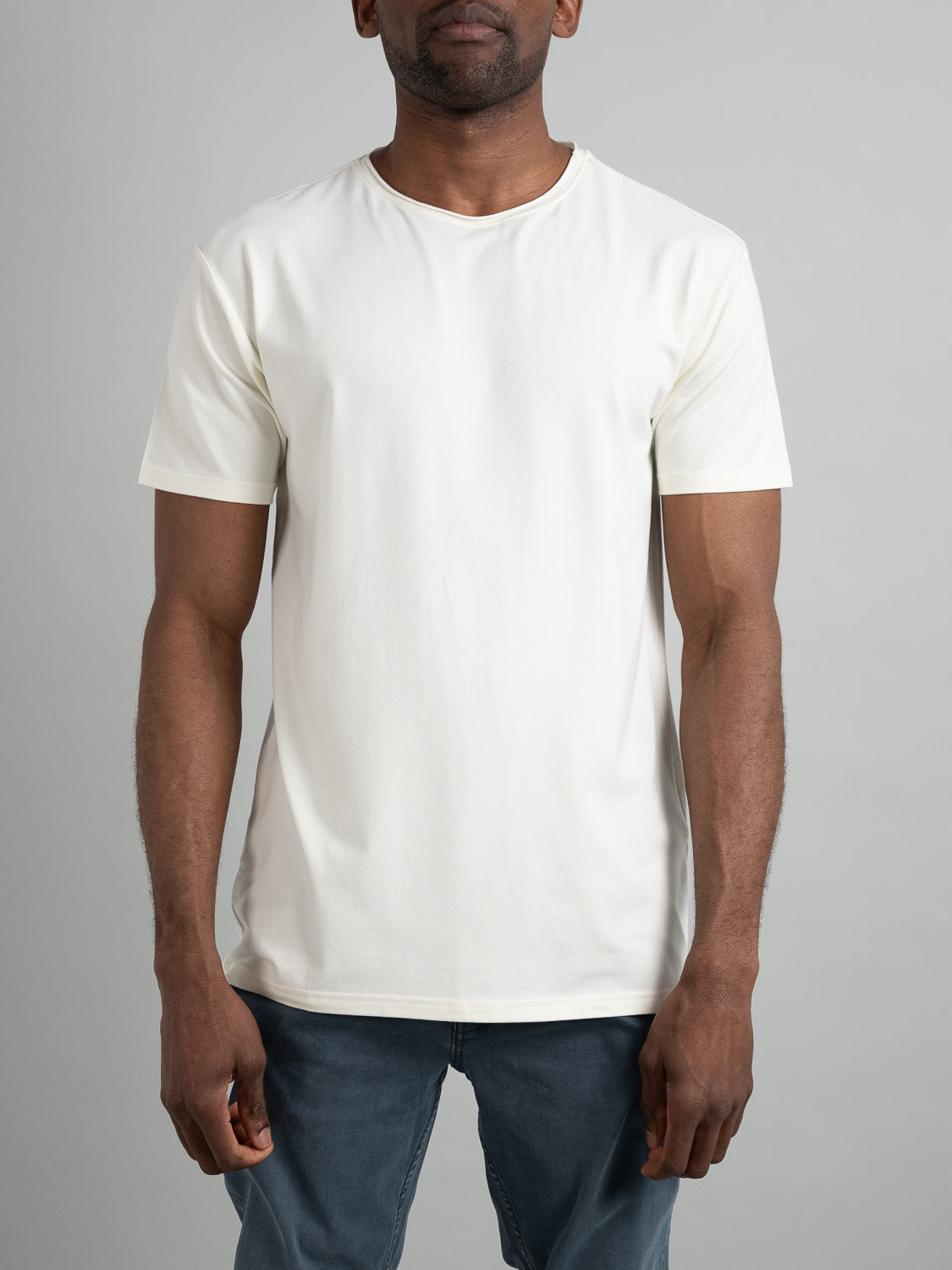 Wearing an ID Menswear Bello Bamboo Blend Rolled Neck Tee and blue jeans, a person stands against a neutral gray backdrop. Their relaxed hands highlight the simplicity of sustainable fashion.