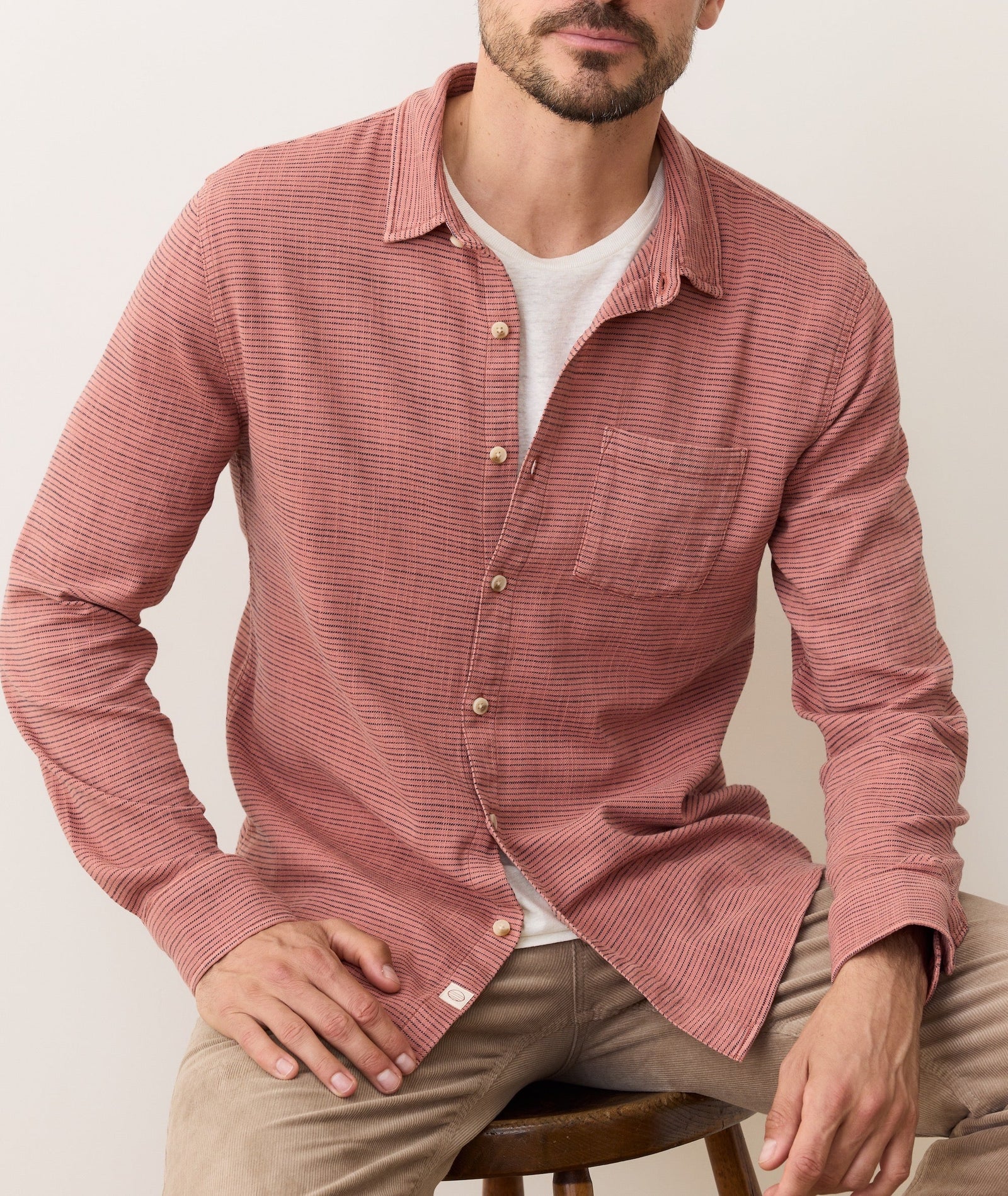 A man sits on a stool wearing Marine Layer's Stretch Selvage Long Sleeve Shirt with a single chest pocket and wooden buttons, layered over a white tee and paired with light brown pants.