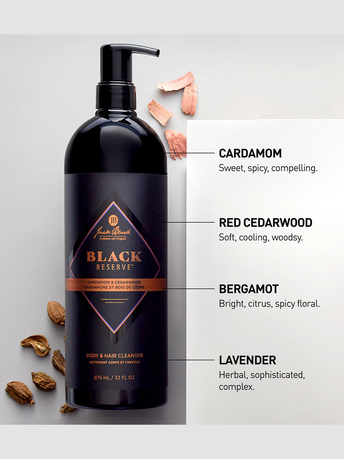 A black bottle of Jack Black Body and Hair Cleanser is displayed with its label visible, next to cardamom pods, cedarwood pieces, and scent notes—highlighting this sulfate-free cleanser made with natural ingredients.