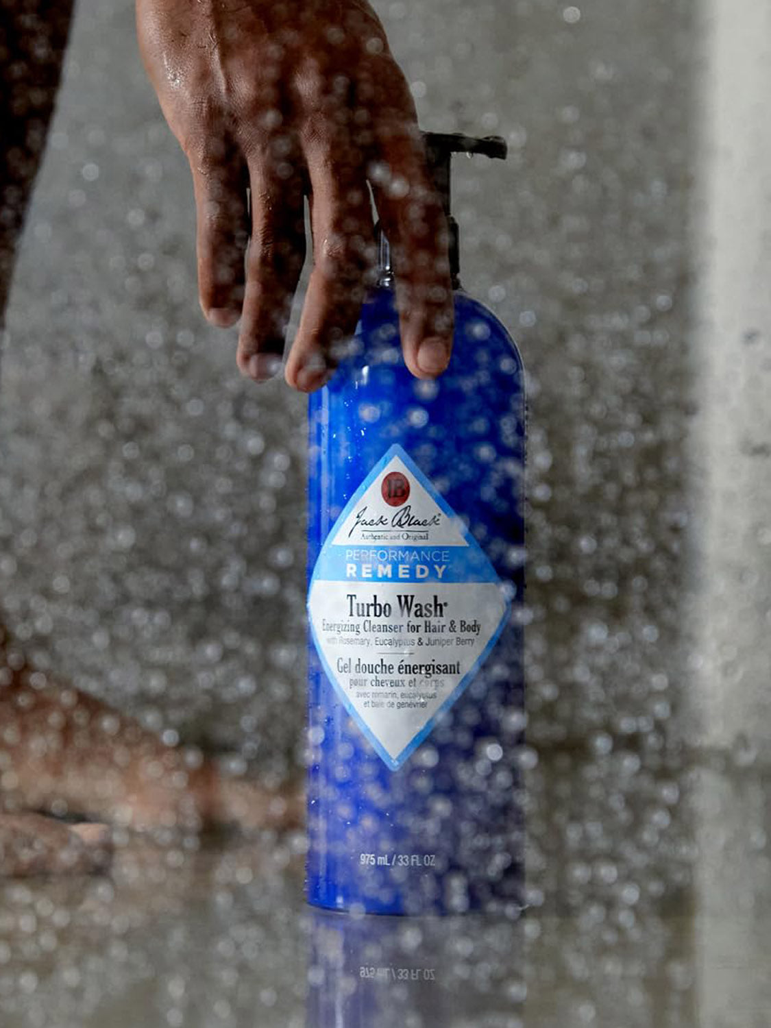 A hand reaches for a blue bottle of Jack Black Body and Hair Cleanser behind a wet, steamy glass shower door dotted with water droplets.