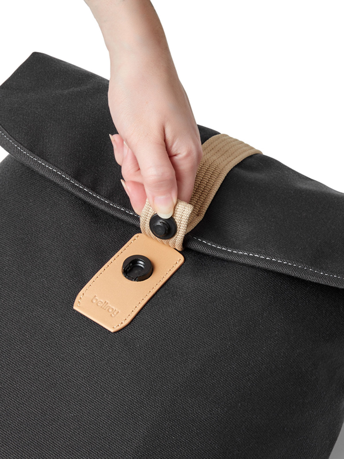 A hand fastens the Bellroy Oslo Backpack—a slim, black fabric minimalist bag with a beige strap and tan leather tag—using a black snap button, set against a white background.