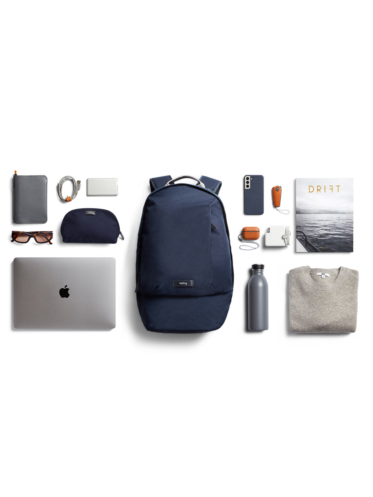 The Bellroy Classic Backpack - 20L in navy blue, made from recycled materials, is displayed with a laptop, gray sweater, sunglasses, notebook, charging cables, water bottle, phone, earphones, magazine, and a matching navy pouch.