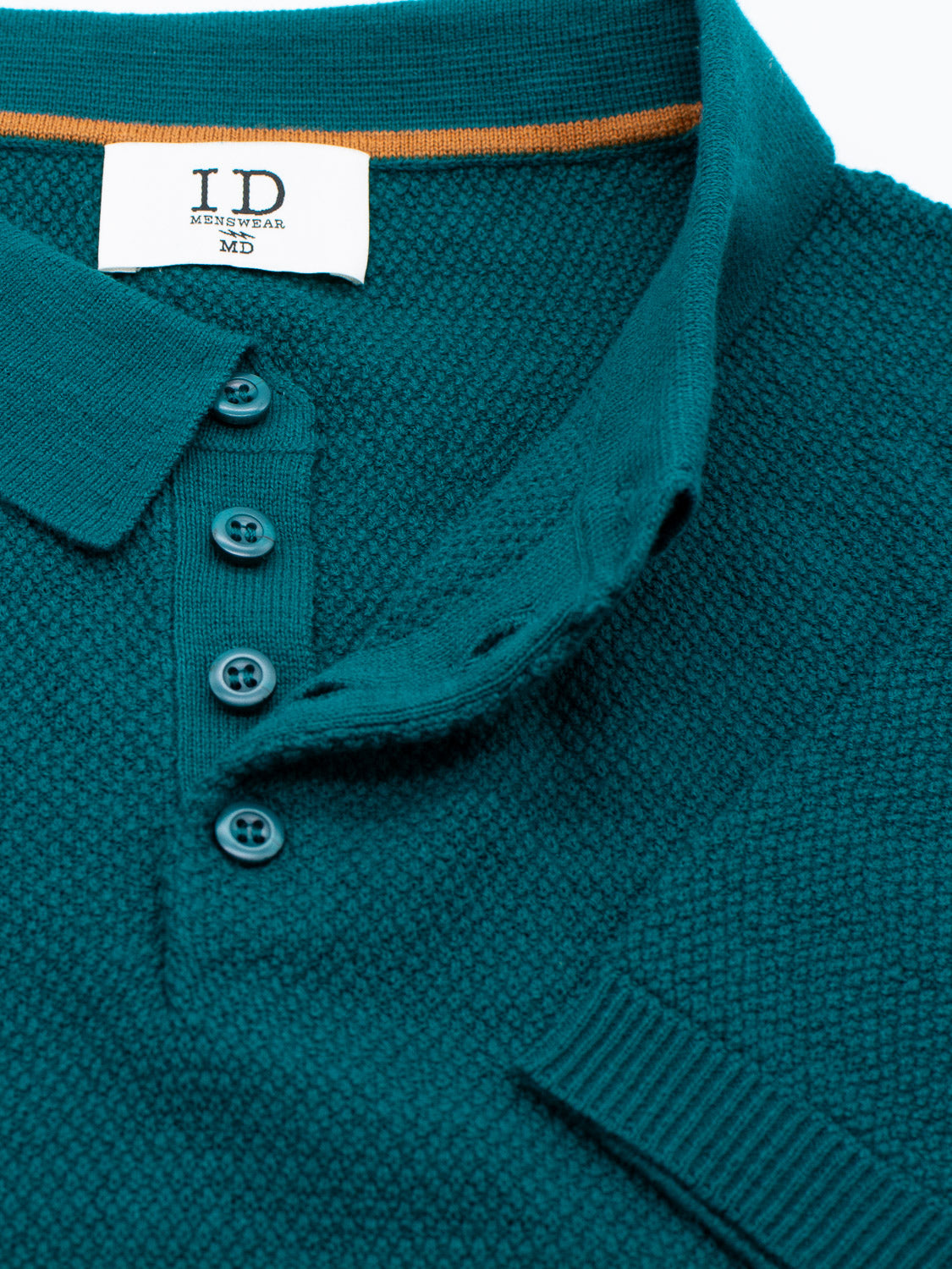 A close-up of the Austin Knitted Cotton Polo from ID Menswear, showcasing a textured teal cotton-knit design with three buttons and a visible brand tag. The breathable fit shirt features an orange stripe inside the collar for contrast.