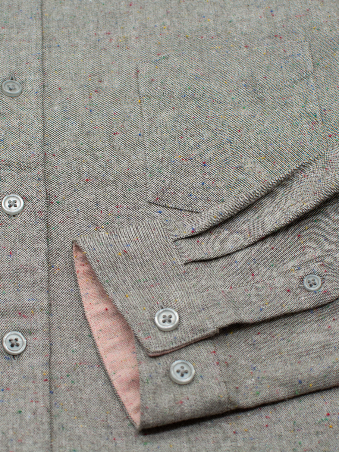 Close-up of the ID Menswear Allswell Speckled Button-Down Shirt showcasing its gray speckled cotton-linen blend, colorful specks, pink cuff lining, four visible gray buttons, and chest pocket.