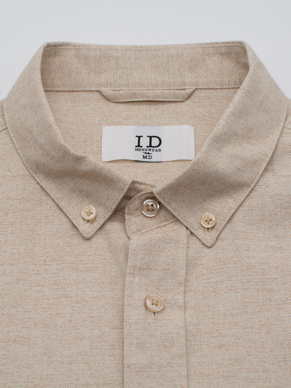 The Maya Fine Herringbone Shirt by ID Menswear is a beige button-down made from 100% cotton with a subtle brushed herringbone pattern, featuring a folded collar and a white tag showing the brand name.