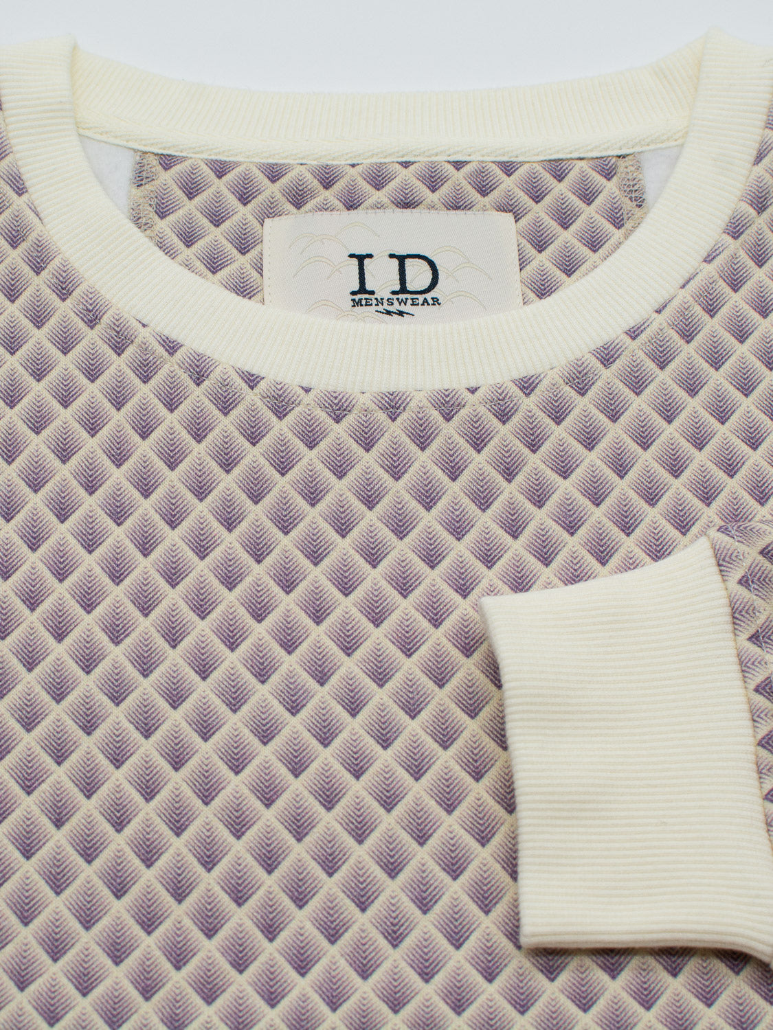 A close-up of the Carp Geometric Printed Sweatshirt reveals a textured, purple and white geometric pattern with cream ribbed edges, exuding a modern artistic vibe. A small right-side pocket is crafted from bamboo and organic cotton fleece. The label reads ID Menswear.