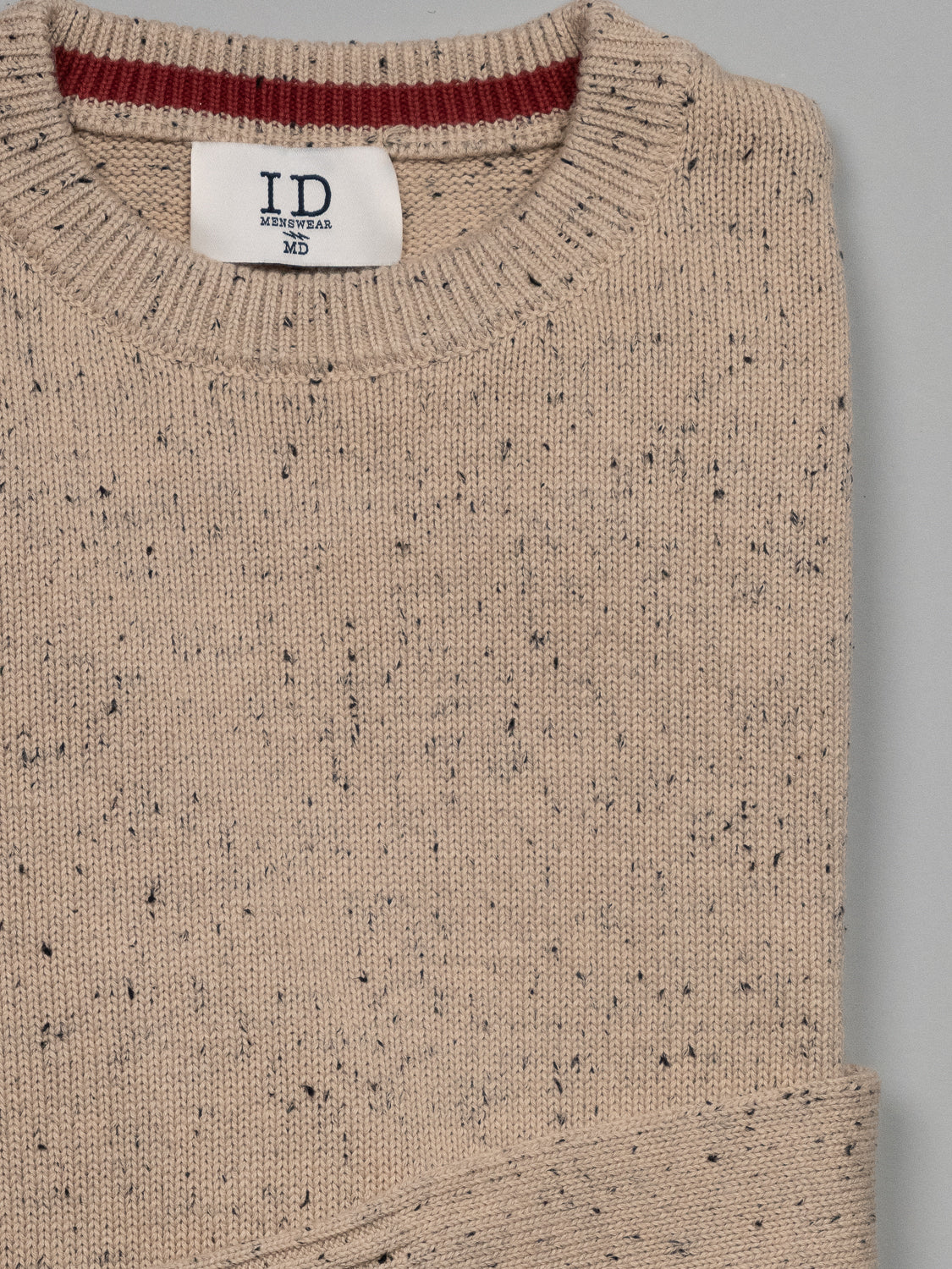 A neatly folded Malmo Speckled Cotton Sweater in beige with dark flecks, featuring an ID Menswear label at the crew neckline, set against a plain light gray background.