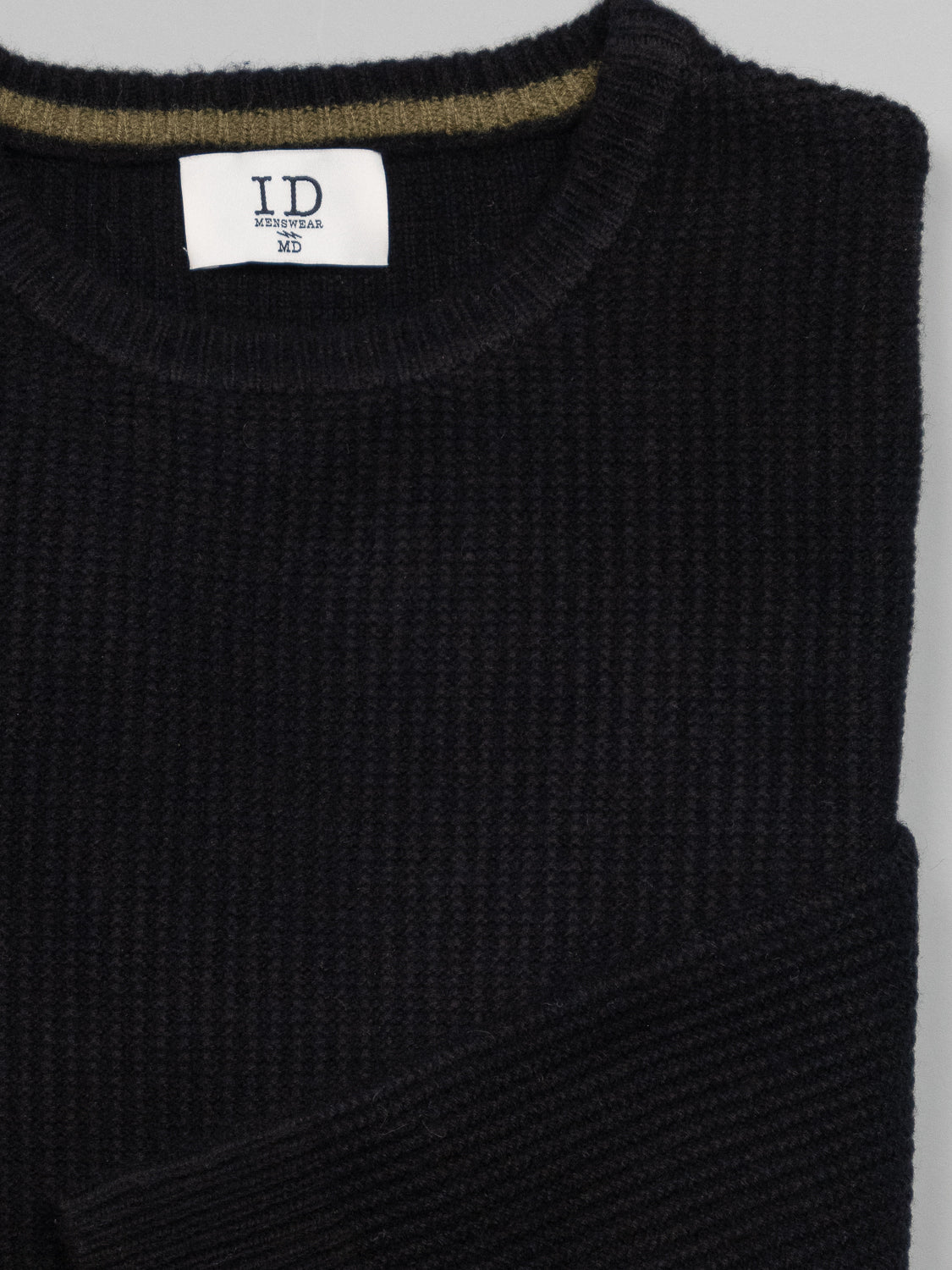 A folded, ribbed black Uppsala Merino Wool Sweater by ID Menswear, featuring a white neck tag, sits neatly against a plain gray background. Perfect for chilly weather.