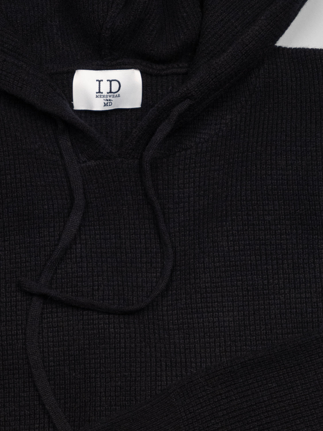 Close-up of the Bergen Waffle Knit Merino Hoody in black by ID Menswear, featuring a ribbed texture and loosely draped hood strings. A visible tag inside the collar reads ID MENSWEAR MD.