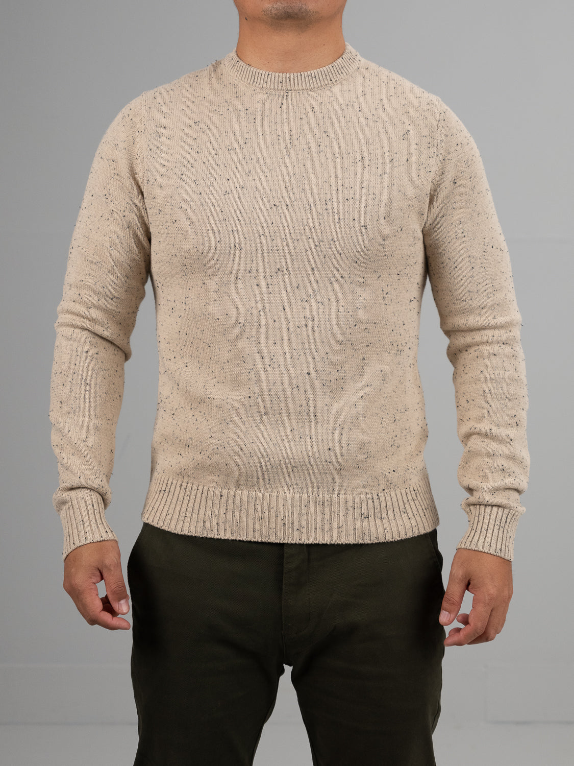 A person, head not visible, stands against a gray background wearing the ID Menswear Malmo Speckled Cotton Sweater in beige with ribbed cuffs and hem, paired with dark green pants.