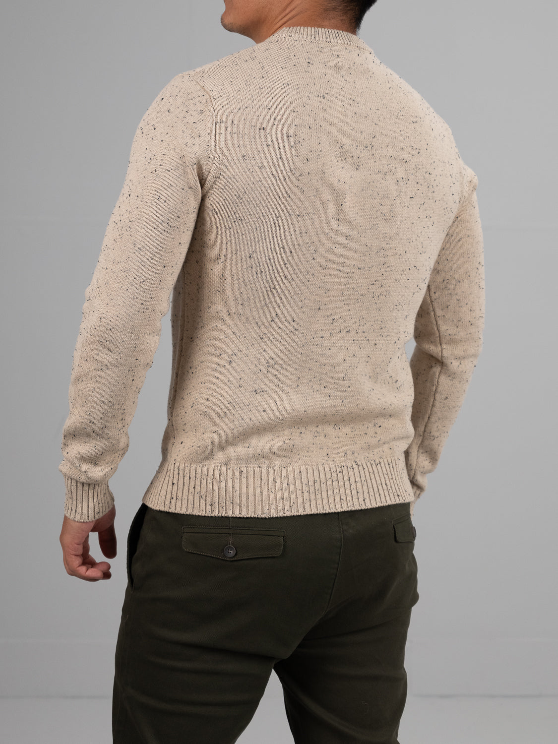A person is seen from the back wearing an ID Menswear Malmo Speckled Cotton Sweater in beige and dark olive green pants, standing against a plain gray background.