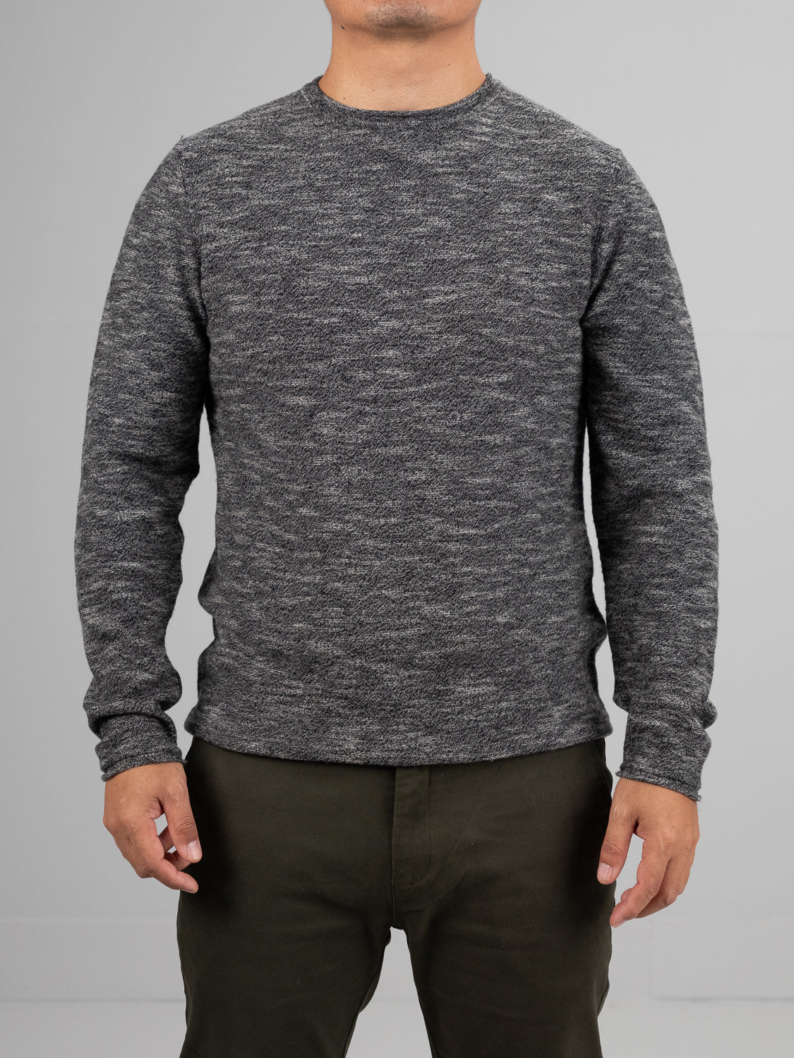 A person stands against a plain gray backdrop wearing ID Menswears Maverick Rolled Neck Sweater, a sophisticated Merino wool design with textured patterns and long sleeves, paired with dark green pants. The lightweight warmth of the sweater blends seamlessly into the scene while their face remains unseen.