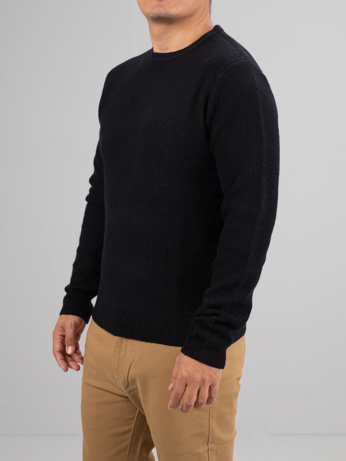 A person in an ID Menswear Uppsala Merino Wool Sweater and light brown pants stands against a plain gray backdrop, facing slightly to the side with arms relaxed by their sides, ideal for chilly weather.