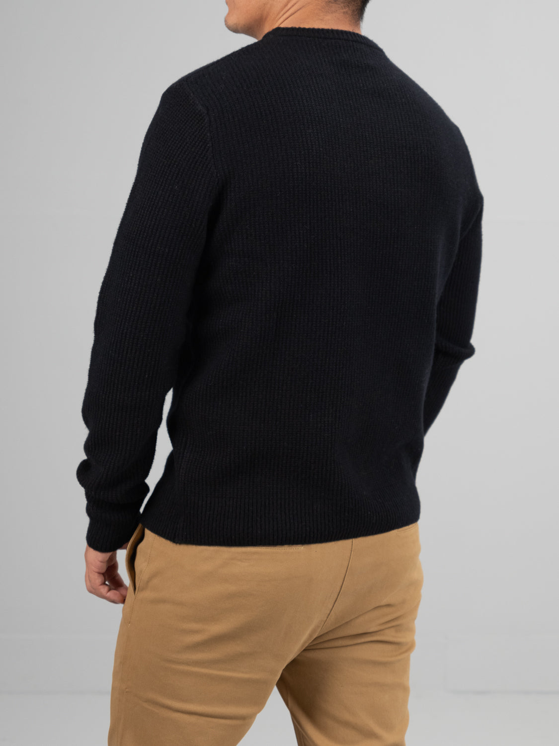A person is wearing an ID Menswear Uppsala Merino Wool Sweater with a ribbed popcorn knit stitch and khaki pants, facing away from the camera against a plain, light-colored background that enhances the chilly weather ambiance.