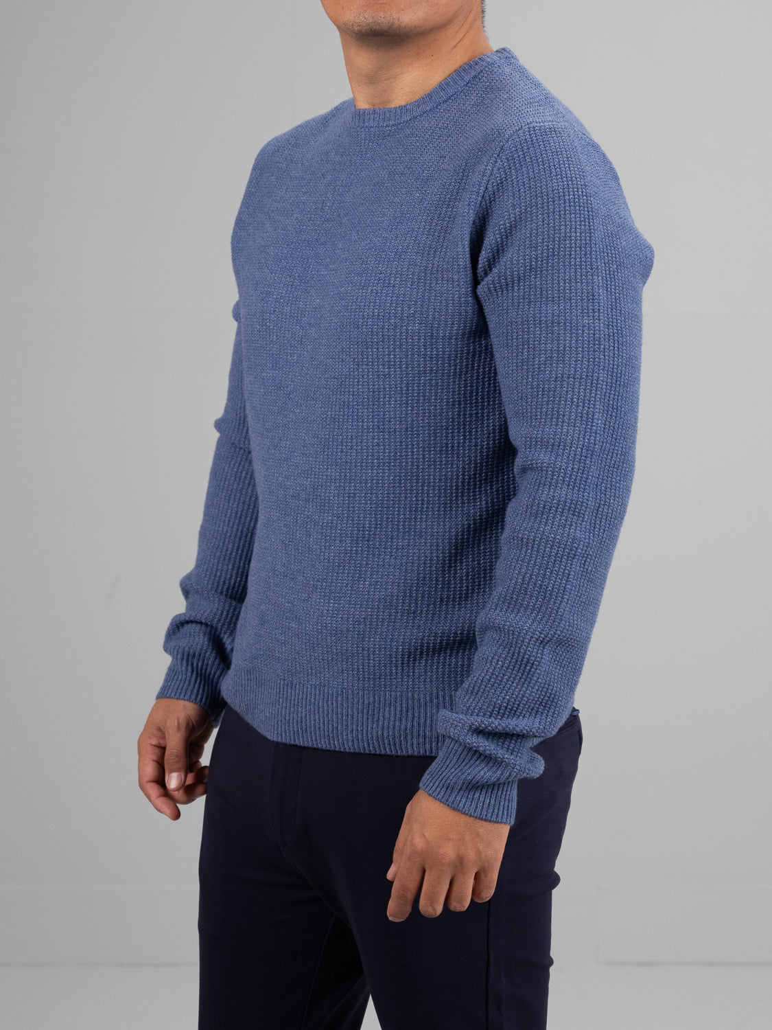 A person wears an Uppsala Merino Wool Sweater by ID Menswear, standing against a light gray background. The blue ribbed popcorn knit highlights cozy style, complemented by dark pants and their relaxed posture, focusing meticulously on the upper body—ideal for chilly weather.