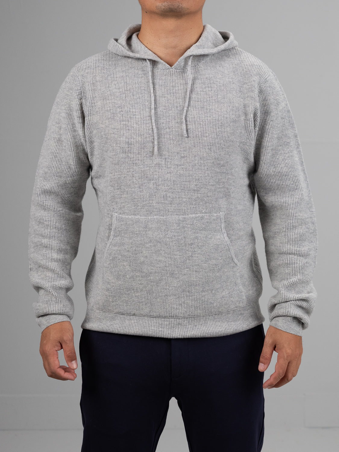 A person wearing the Bergen Waffle Knit Merino Hoody by ID Menswear, featuring a light gray color, front pocket, and loose drawstrings. The background is plain, and the persons face isn’t visible.