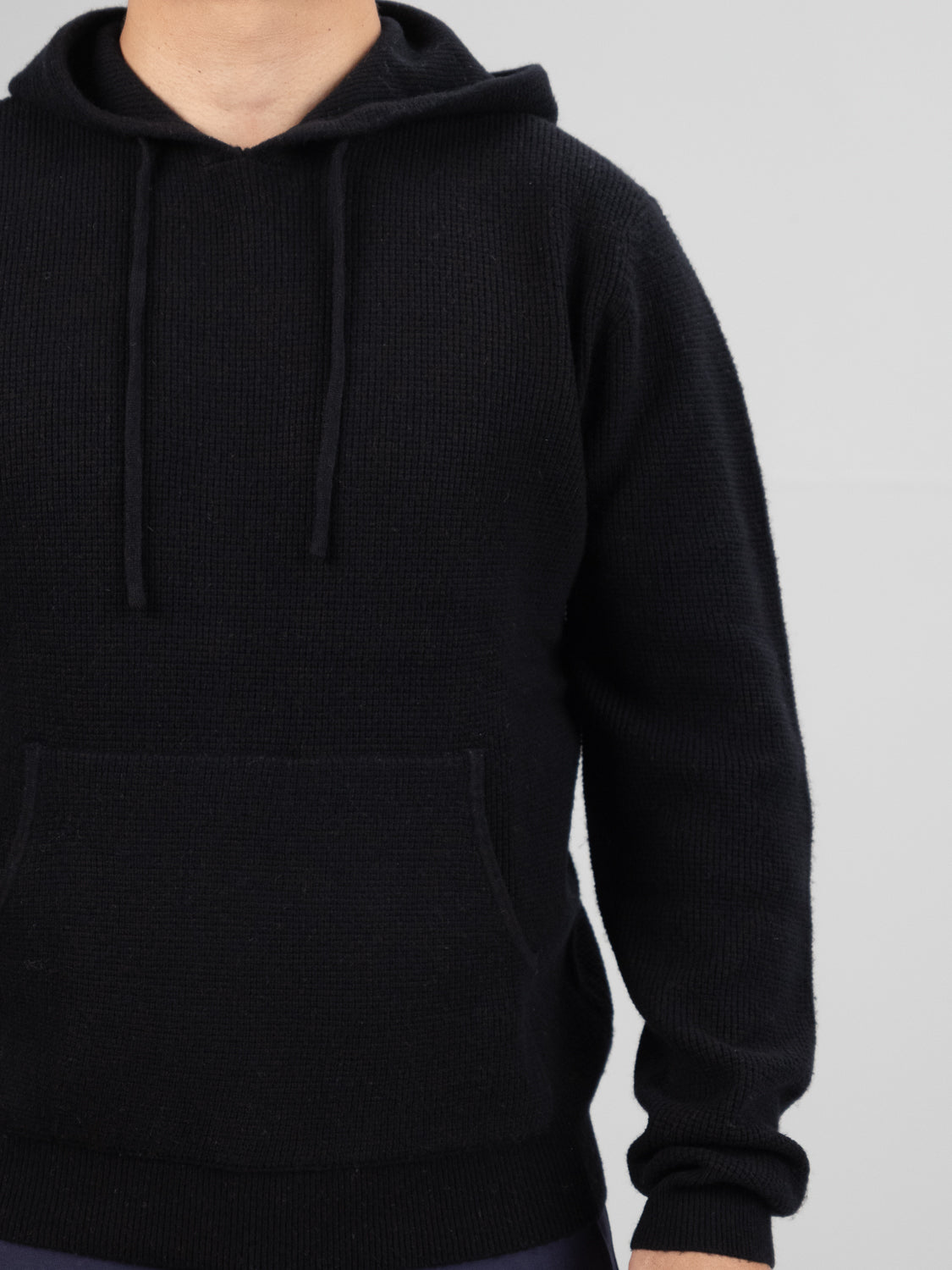 A person wears the Bergen Waffle Knit Merino Hoody by ID Menswear. The black hoodie, featuring a front pocket and drawstrings, is set against a plain gray background. The image is cropped above the shoulder, leaving the face unseen.