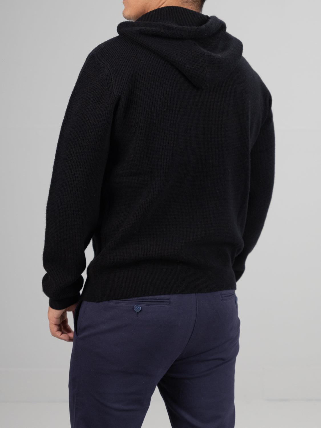 A person in an ID Menswear Bergen Waffle Knit Merino Hoody, black in color, and dark blue pants stands with their back facing the camera against a plain light-gray backdrop.