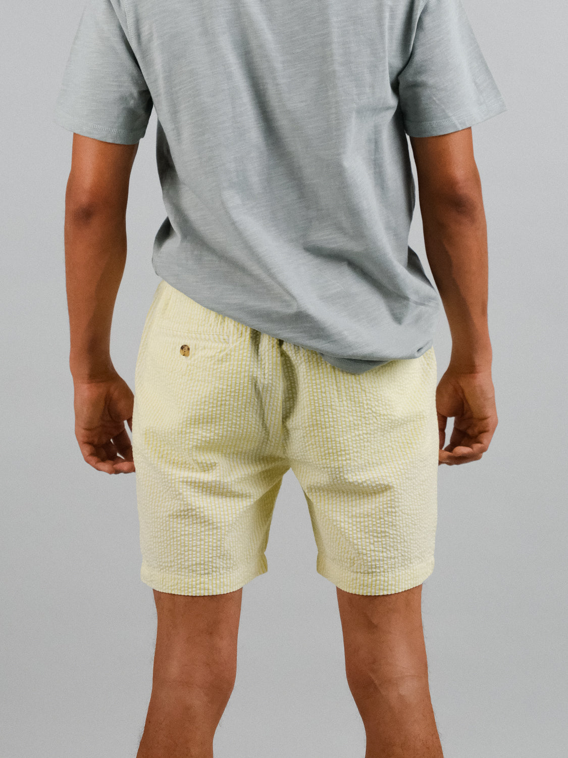 A person faces away, wearing a light gray t-shirt paired with yellow Ibiza Seersucker Drawstring Shorts by ID Menswear. The plain, neutral background highlights this casual ensemble, ideal for warm weather wardrobes.