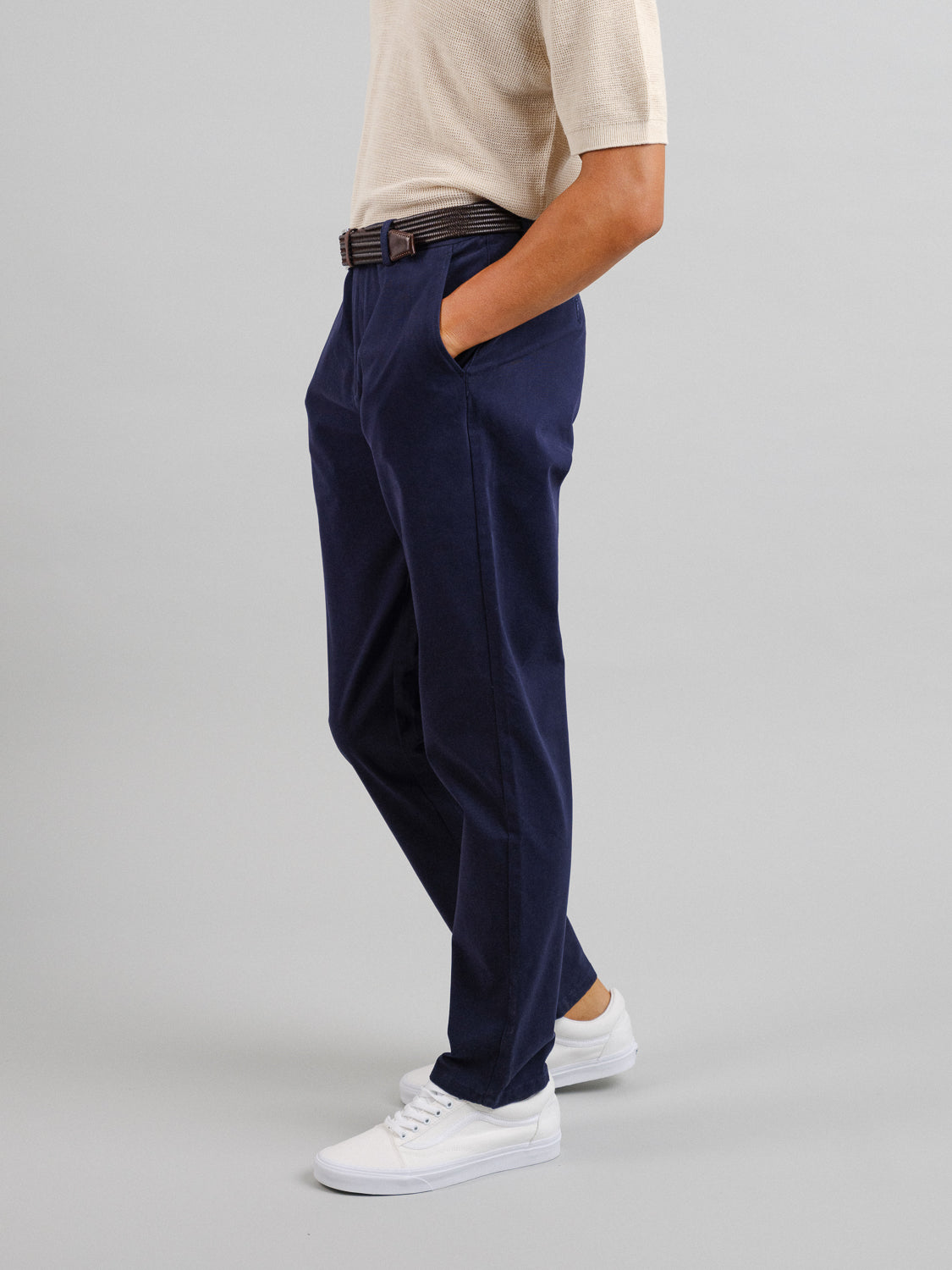 A person in a beige short-sleeve shirt tucked into relaxed-fit Bushwick Twill Stretch Chino trousers by ID Menswear, with hands in pockets, paired with a brown belt and white sneakers, stands against a plain gray background.