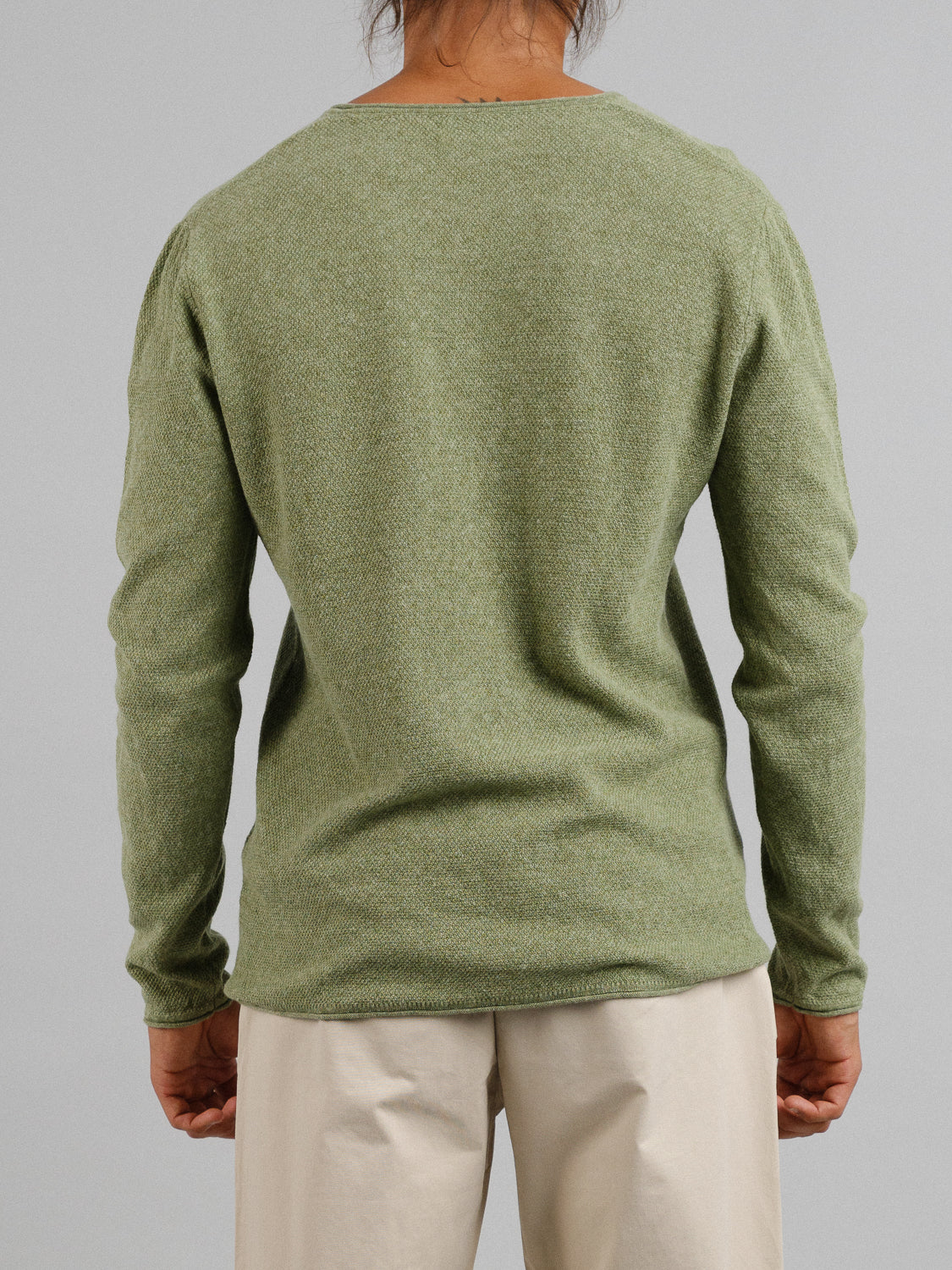 Against a plain gray background, a person with long hair turns their back to the camera, beautifully highlighting an Amalfi Deep V Knitted Top from ID Menswear in light green linen-cotton blend, stylishly paired with beige pants.