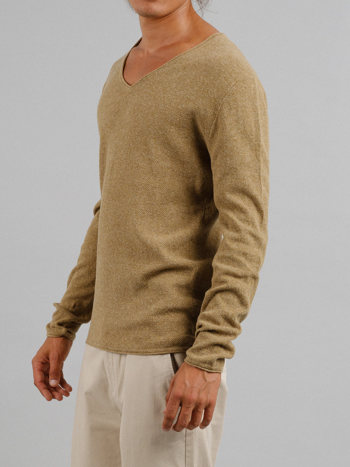 A model is shown from the shoulders down, slightly turned to the side, wearing ID Menswears Amalfi Deep V Knitted Top in light brown paired with beige pants against a plain gray background.