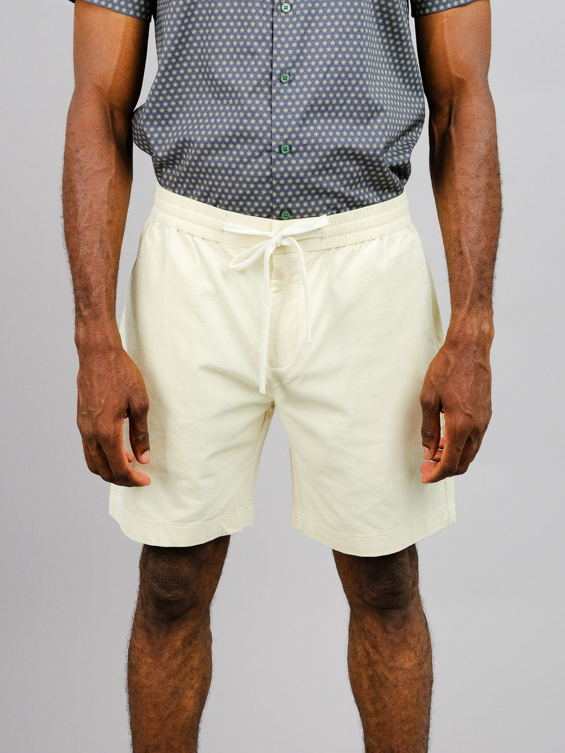 A person models an ID Menswear patterned short-sleeve shirt with relaxed-fit Odyssey Corduroy Stretch Shorts in light beige. The elastic waistband shorts stand against a plain gray wall backdrop, with the crop framing them from the shoulders down.