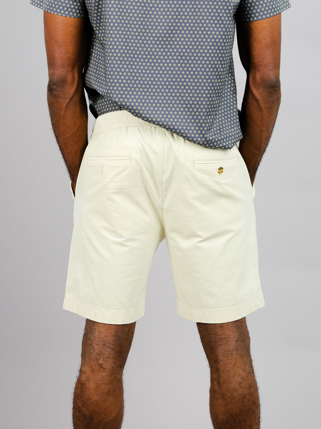 A person models ID Menswears Odyssey Corduroy Stretch Short in light beige, paired with a grey patterned shirt. These relaxed fit shorts feature an elastic waistband and two buttoned back pockets, set against a plain gray backdrop.