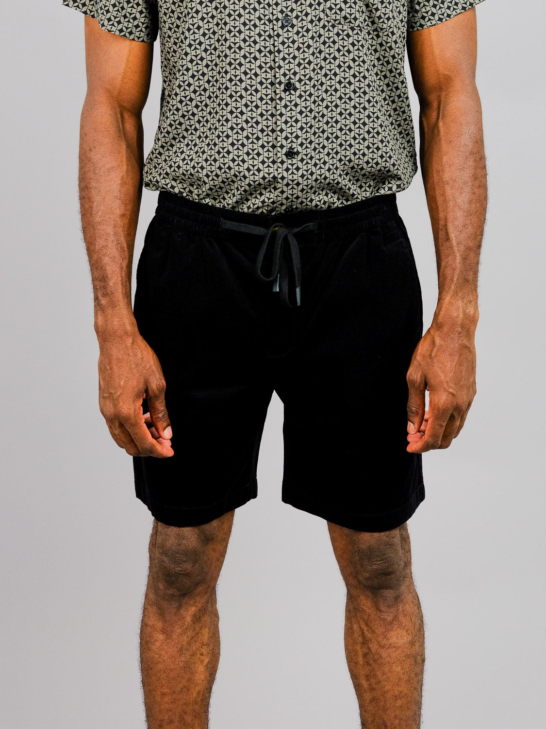 A person stands against a plain background wearing the Odyssey Corduroy Stretch Short by ID Menswear. These black, elastic-waist drawstring shorts feature a patterned short-sleeve shirt and offer a relaxed above-the-knee fit. Only the upper legs to the waist are visible.