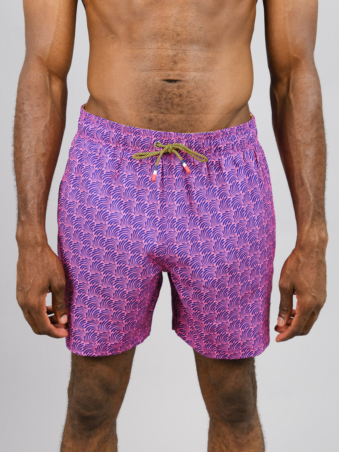 Against a plain background, a person stands wearing ID Menswears Nazare Fast Dry 4-Way Stretch Swim Trunks in vibrant pink. Crafted from recycled plastic bottles with a wave-like pattern and drawstring, their torso and legs are visible, with hands relaxed at their sides.