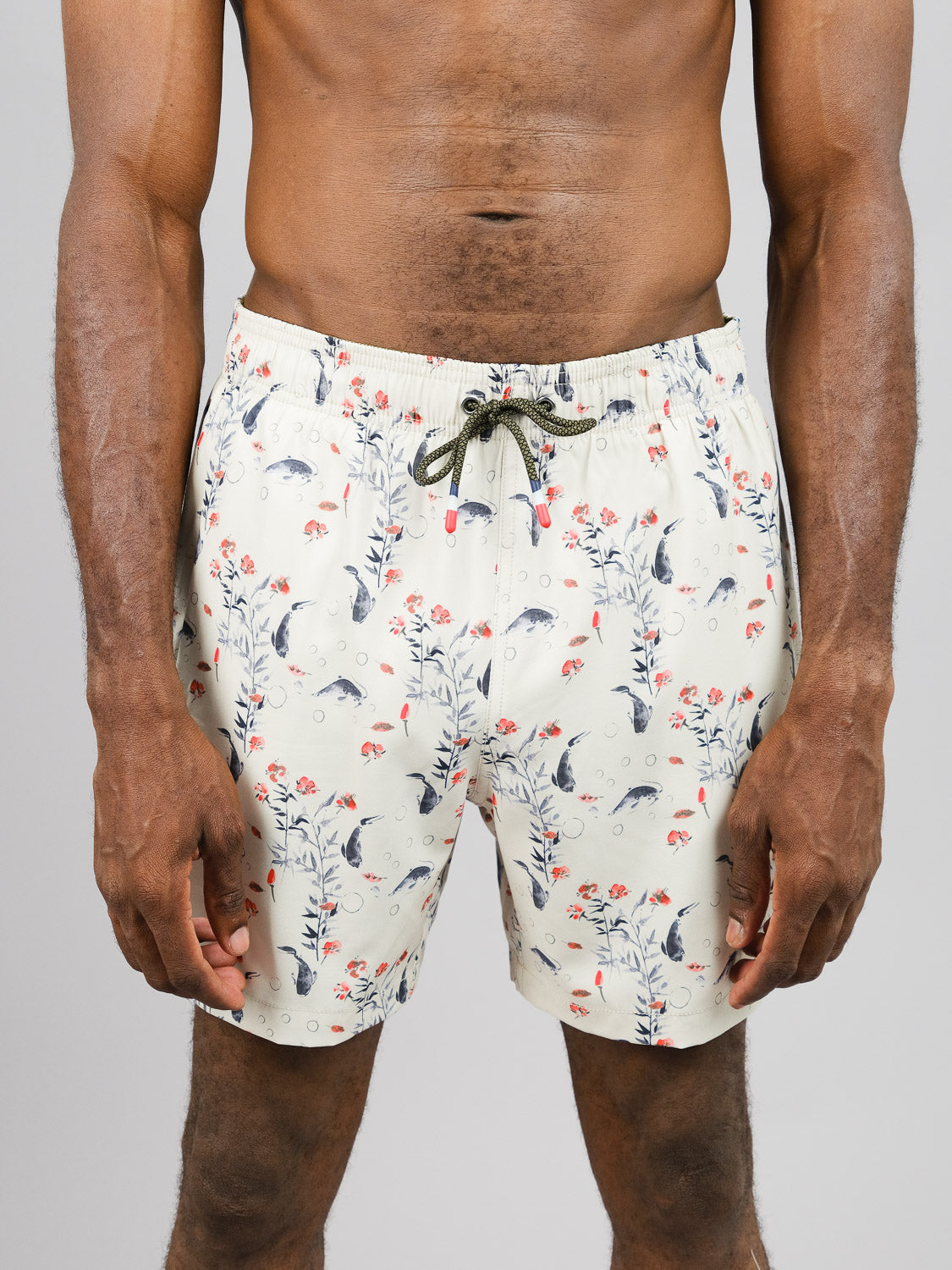 Someone is wearing the Koi Fast Dry 4-Way Stretch Swim Trunk from ID Menswear, featuring a light-colored floral and fish pattern made from recycled bottles. With a green drawstring waist and quick-dry fabric, it ensures comfort against a neutral background.