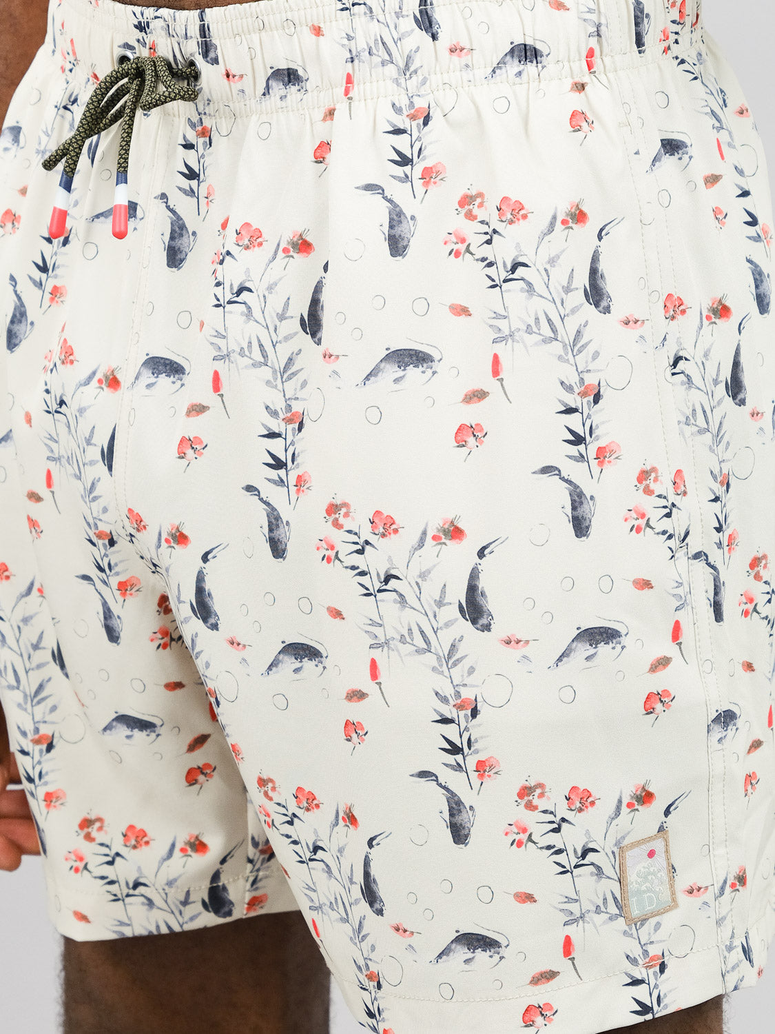 Close-up of a person wearing ID Menswear’s Koi Fast Dry 4-Way Stretch Swim Trunk—cream shorts with a koi fish, pink flower, green leaf, and bubble pattern. Features quick-dry fabric and an elastic waistband with green and gray drawstring.
