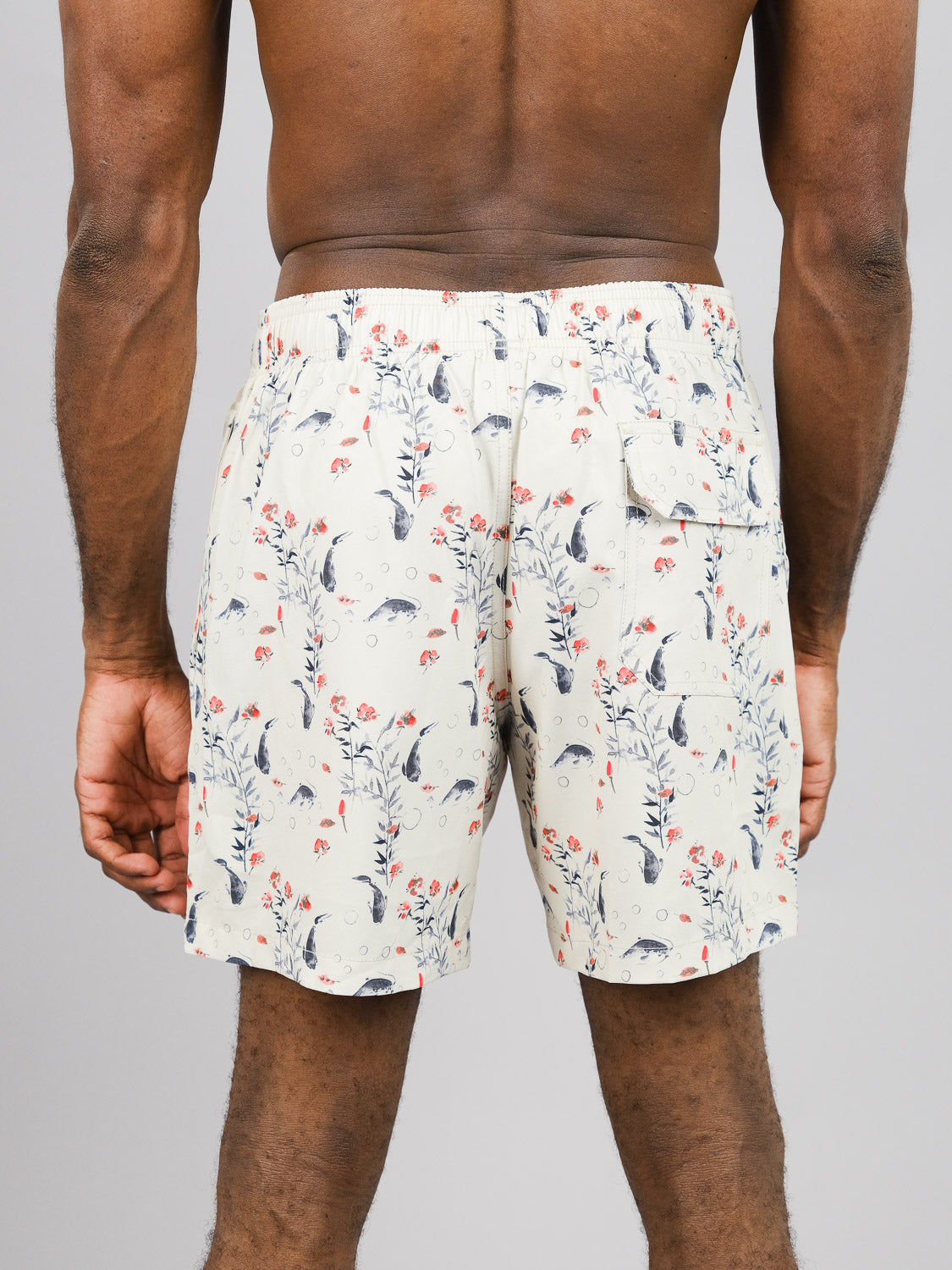 The back view shows a person wearing ID Menswears Koi Fast Dry 4-Way Stretch Swim Trunk, featuring a nature-themed pattern of leaves, flowers, and small animals. The light-colored shorts are made from recycled plastic bottles with an elastic waistband and a back pocket against a gray background.