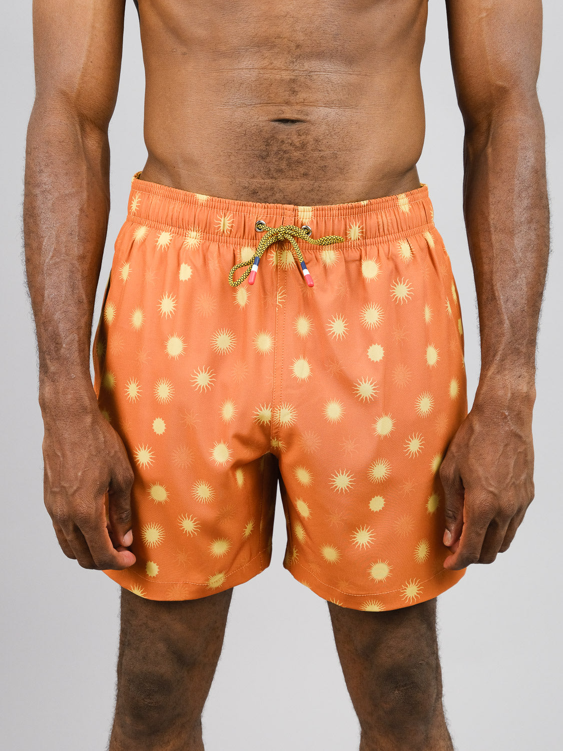 Wearing Sol Fast Dry 4-Way Stretch Swim Trunk by ID Menswear, a person showcases quick-dry orange swim shorts with a yellow sunburst pattern. Made from recycled plastic bottles, the shorts have a green drawstring with red and blue tips. Theyre posed against a plain gray background showing torso and legs only.